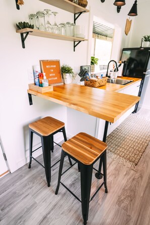 A kitchen with a wooden countertop, two stools, a shelf with glassware, and a welcome sign.