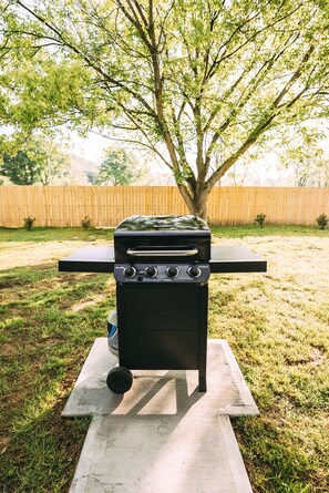 Ein tragbares Grillset auf einer Betonplatte in einem Hinterhof mit Holzzaun und Bäumen im Hintergrund.