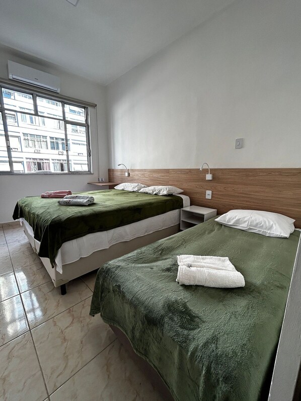 A hotel room with two beds, a window with a view of buildings, and a tiled floor.
