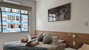 Een slaapkamer met een bed, nachtkastje, lamp, raam met uitzicht op gebouwen en een schilderij aan de muur.