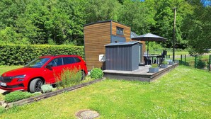 A red SUV parked in a grassy area with a small wooden structure and a patio with outdoor furniture. - Tiny Haus Tinyhouse Waldcamping Prüm Eifel (Prüm)