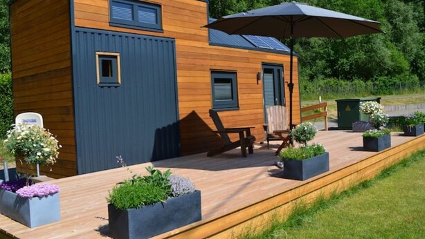 A modern wooden cabin with a deck, outdoor seating, and a large umbrella. - Tiny Haus Tinyhouse Waldcamping Prüm Eifel (Prüm)