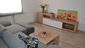 Un salon moderne avec un canapé gris, une table basse en bois ornée d’un vase et de deux verres, et un meuble TV en bois sur lequel sont posés un vase à fleurs et deux tableaux.