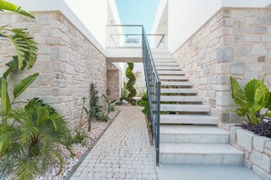 Un escalier moderne, avec des murs en pierre et une rampe en métal, entouré de plantes en pot et bordé d’un chemin pavé.