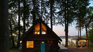 Una cabaña de madera con puerta azul, rodeada de altos árboles y vista al lago.