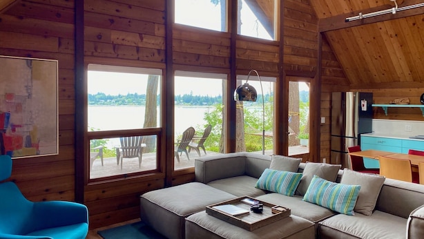 Un interior acogedor de cabaña con un gran techo de madera, un conjunto de sofás, una mesa de comedor y vistas a un lago a través de amplios ventanales.