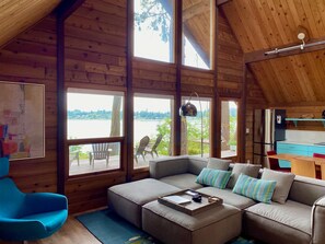 A cozy cabin interior with a large wooden ceiling, a sofa set, a dining table, and a view of a lake through large windows. - Waterfront Burton Loop - The Very Best Of Vashon, cozy cabin with a harbor view. (Vashon)