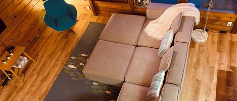 Ein gemütliches Zimmer mit einer Eckcouch, einem blauen Sessel, einer runden Lampe und Holzboden.