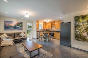 Una sala moderna con un sofá, una mesa de centro y obras de arte. Junto a ella, una cocina con armarios de madera, un refrigerador y un comedor.
