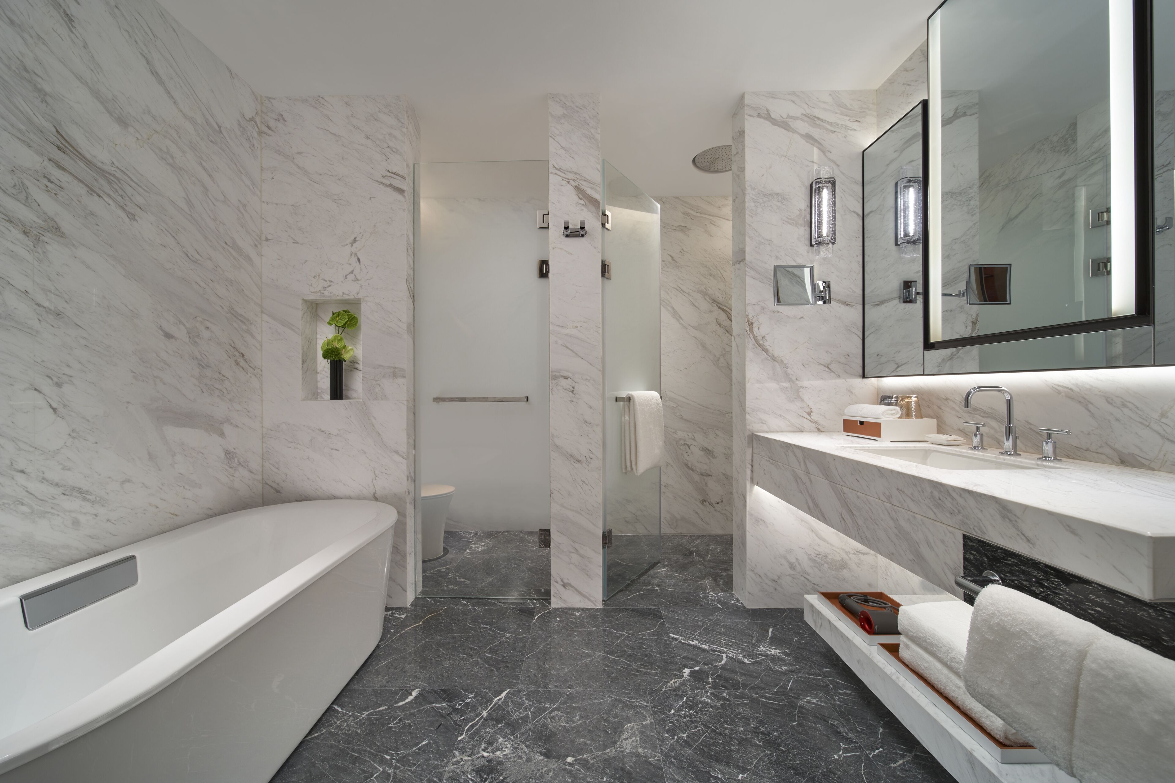 A modern bathroom with a marble wall, a bathtub, a vanity with a mirror, and a shower area.