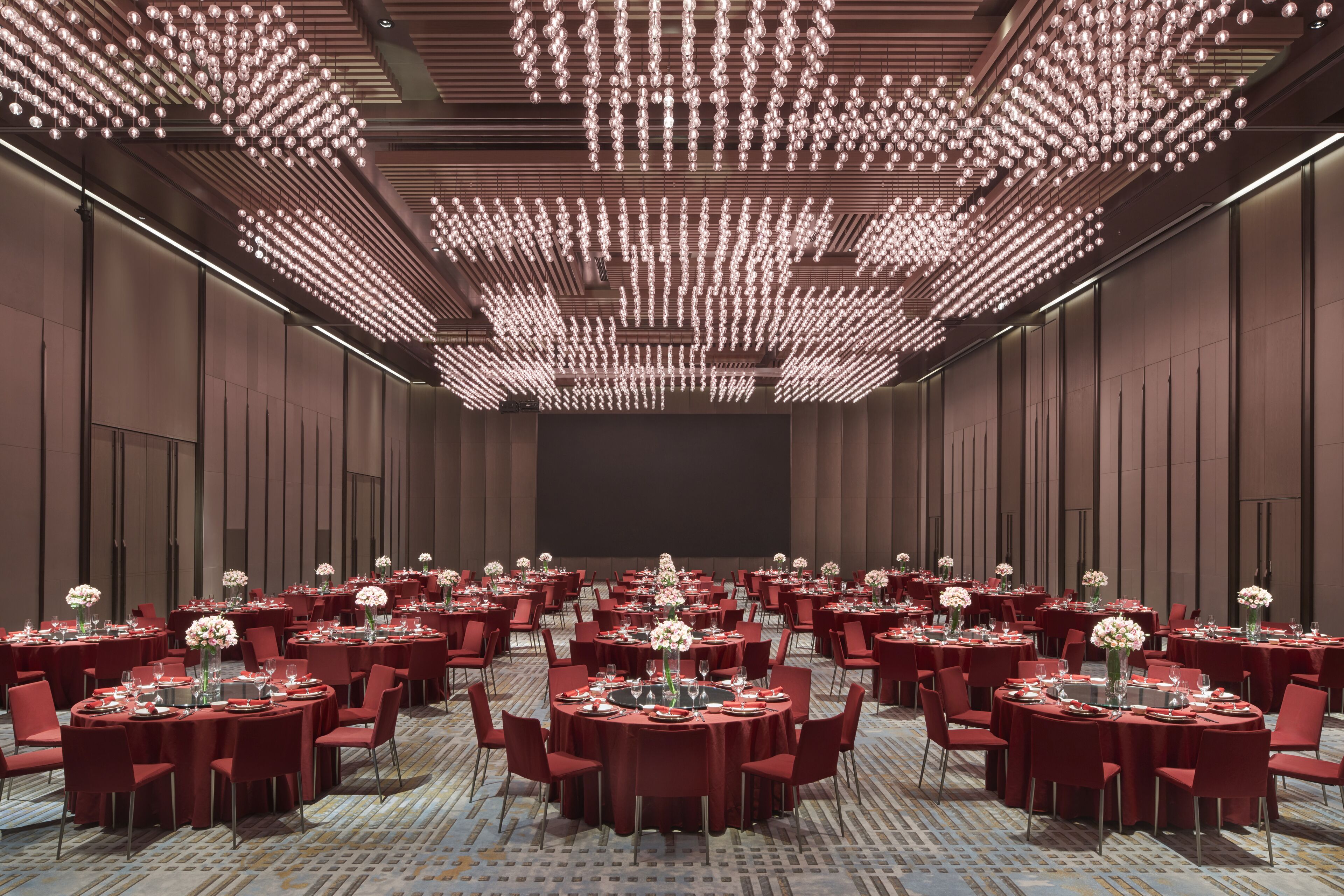 A large conference room with multiple round tables, each set with red tablecloths, chairs, and floral centerpieces.