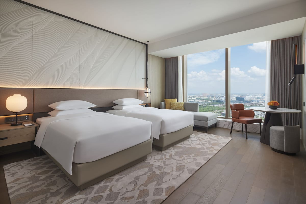 Une chambre d'hôtel moderne avec deux lits, un coin salon avec une table et des chaises, une grande fenêtre donnant sur la ville et un mur décoré de motifs.
