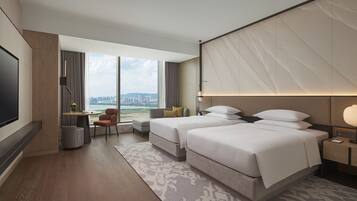 A modern hotel room with two beds, a TV, a desk, and a large window offering a city view.