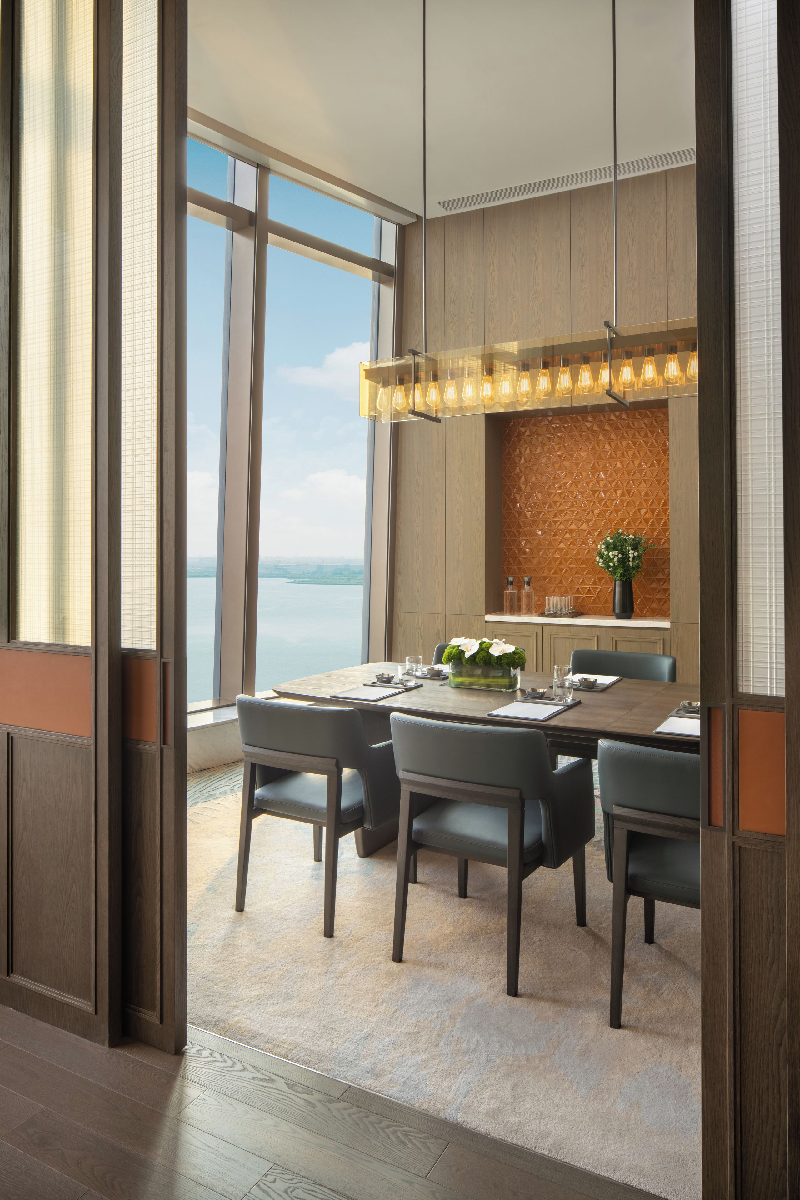 A modern dining area with a view of the sea, a wooden wall with geometric patterns, and a chandelier.