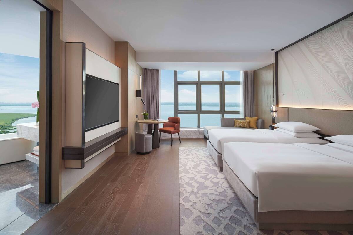 Une chambre d’hôtel avec une grande télévision, deux lits, une petite table avec une chaise, et une vue sur la mer.