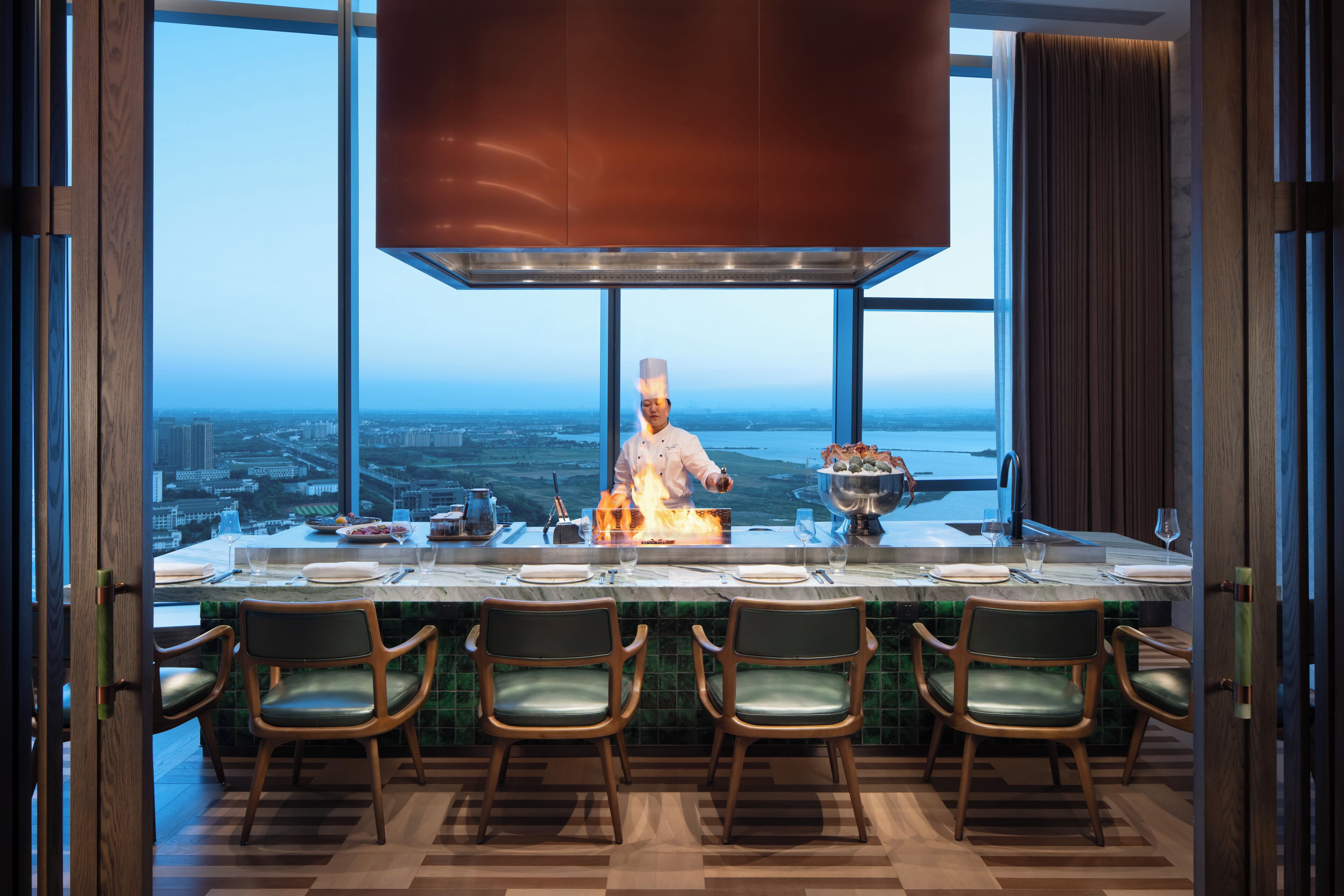 A chef cooking at a high table with a city view, surrounded by chairs and a large window.