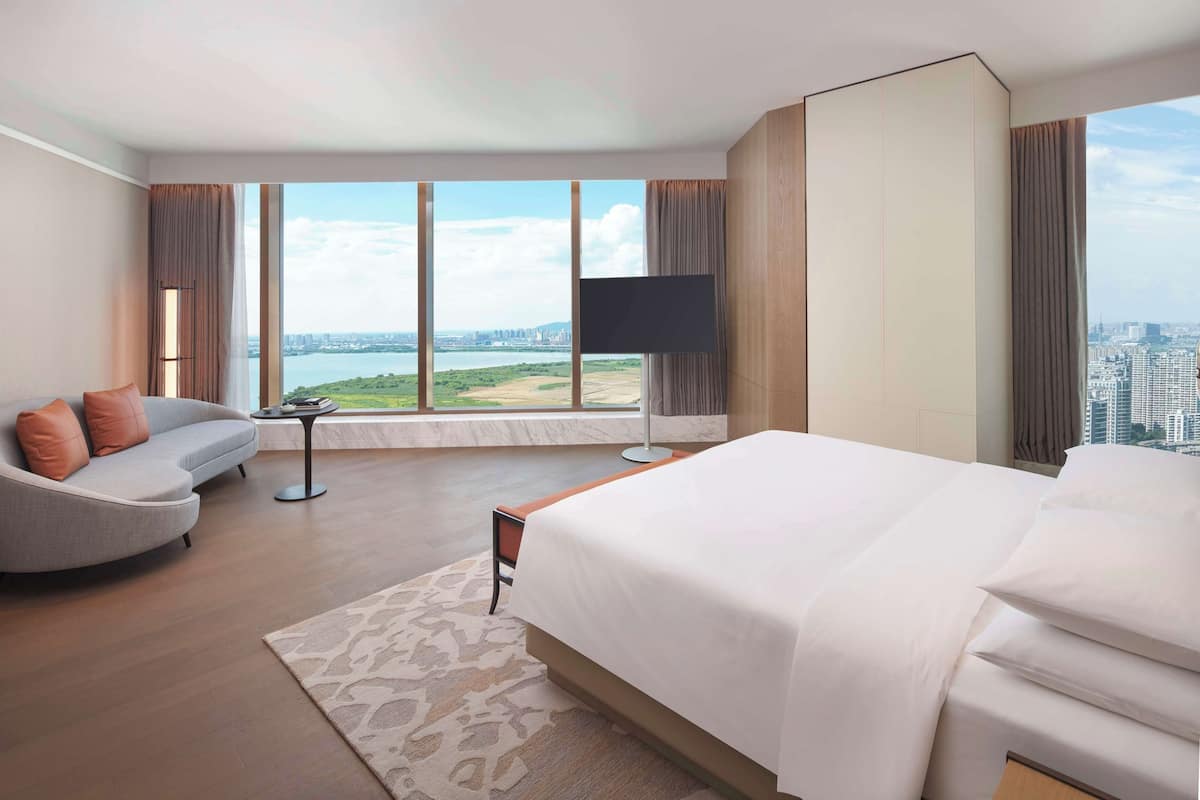 Une chambre d’hôtel moderne dotée d’un grand lit, d’un canapé, d’une télévision et offrant une vue sur le paysage urbain et l’eau.