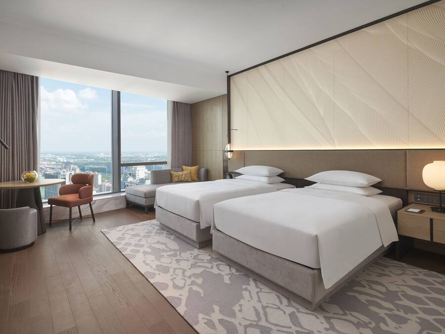 Quarto de hotel moderno com duas camas, um espaço de estar pequeno e uma janela grande com vista para a cidade.