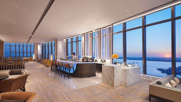 A spacious hotel lobby with a bar, seating area, and large windows offering a view of the sunset over the water. - Hyatt Regency Changshu Kuncheng Lake (Suzhou)