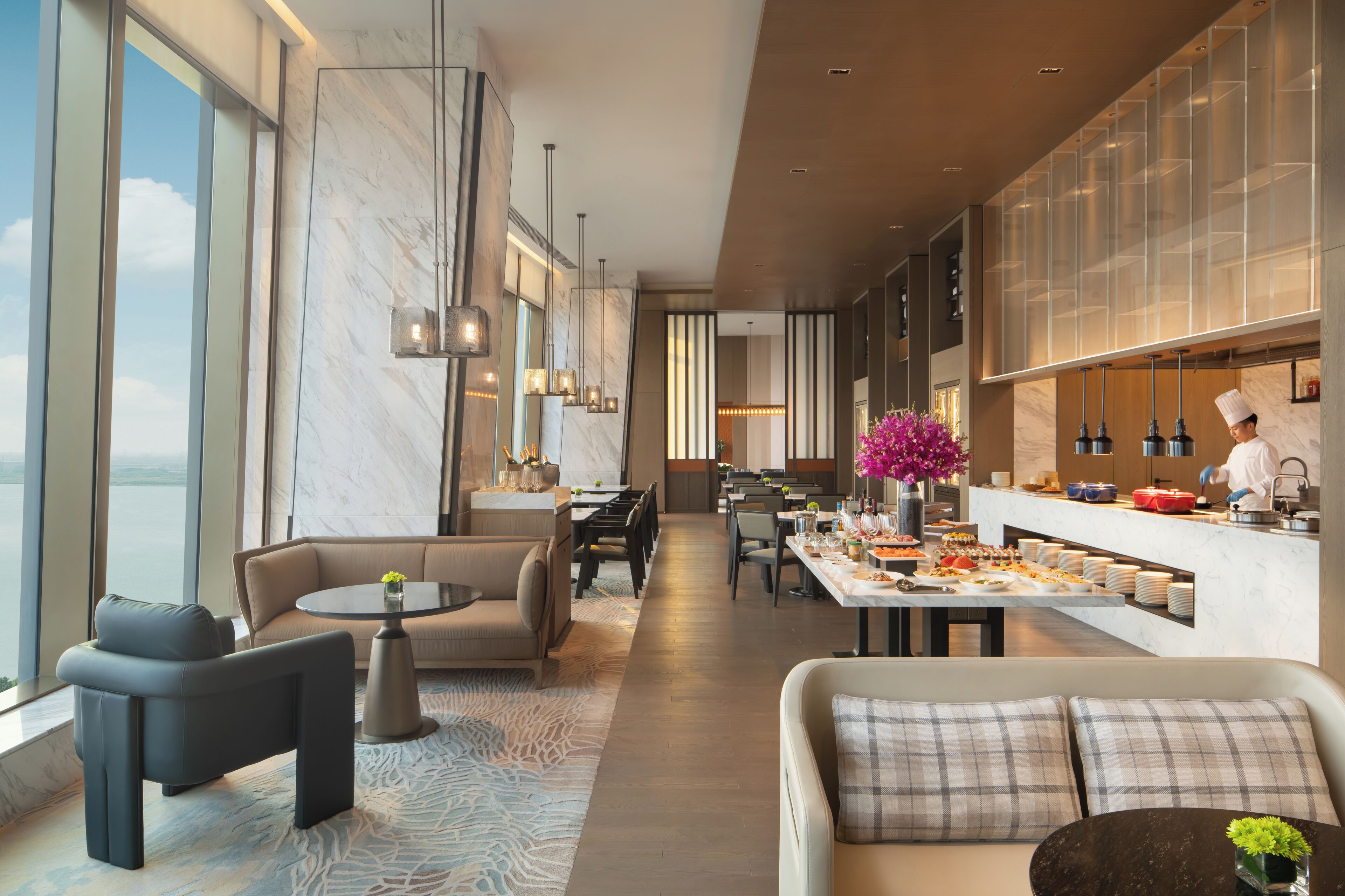 A modern hotel lobby with a dining area, seating arrangements, and a kitchen visible in the background.
