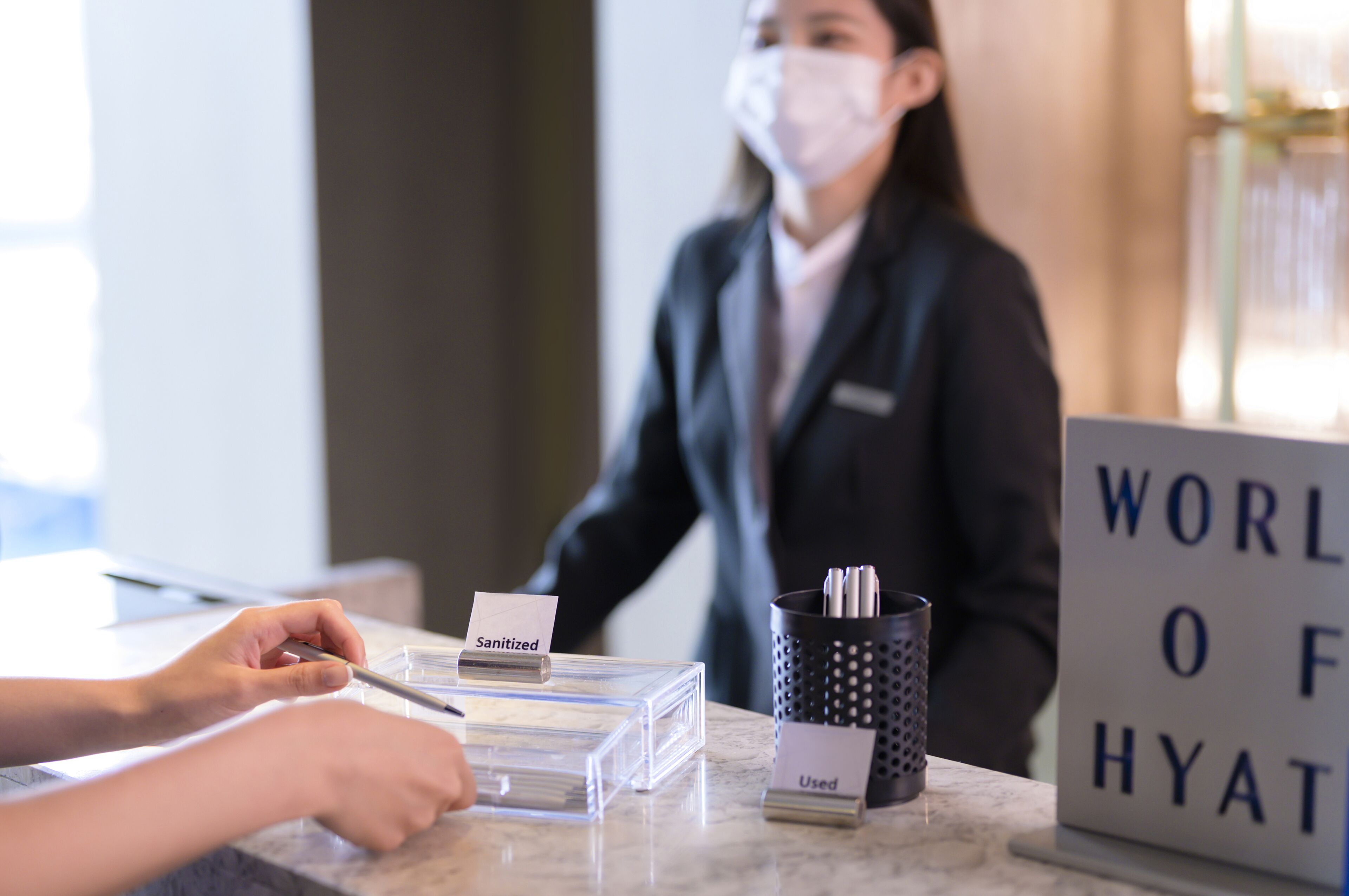A hotel reception desk with a sign reading "WORLD OF HYAT". There are two clear plastic boxes labeled "Sanitized" and "Used". A person is standing behind the desk, and there is a black perforated container with pens.