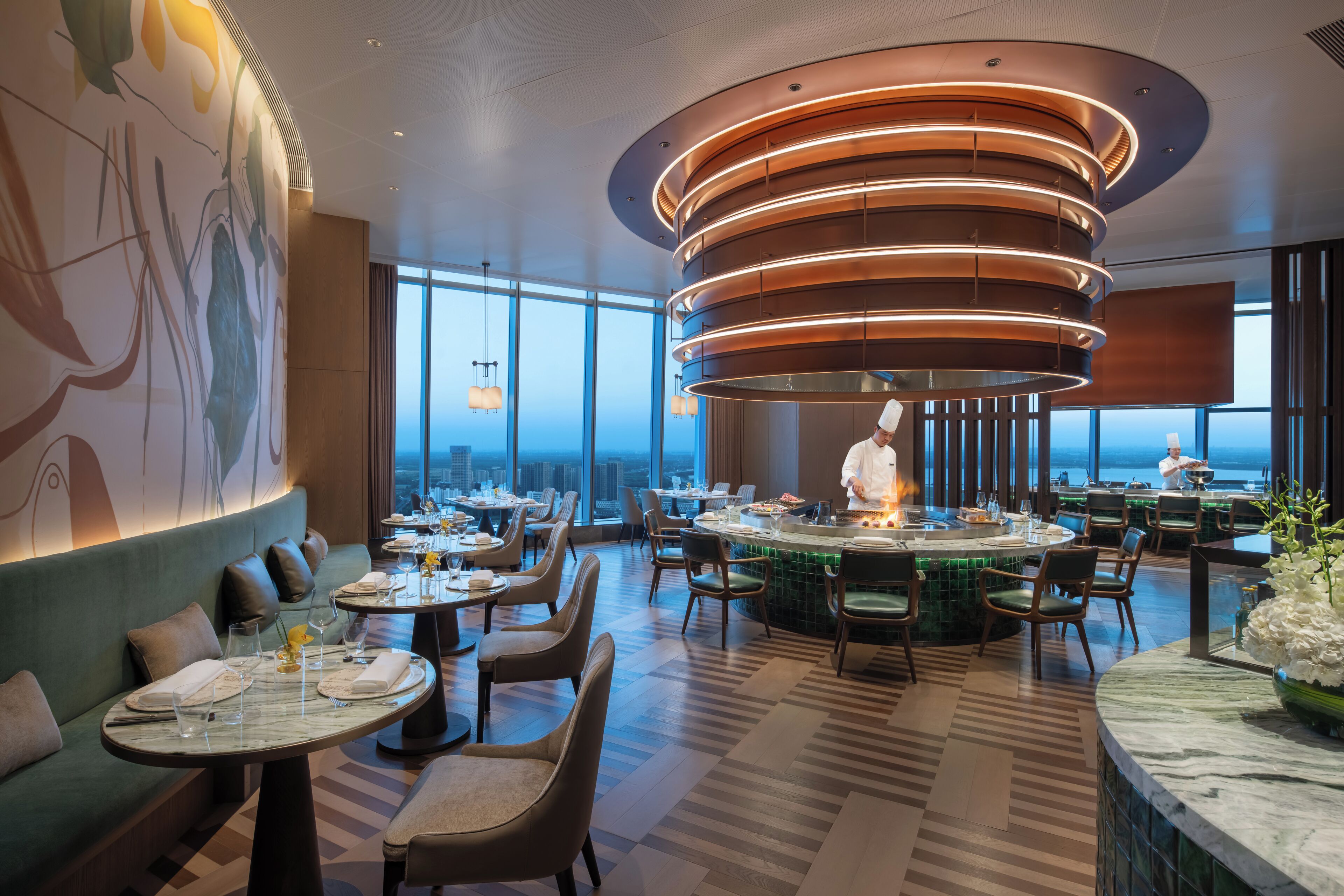 Un restaurante moderno con una isla central, techos altos y amplios ventanales con vistas a la ciudad.