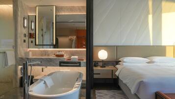 A modern hotel room with a large bathtub, a separate shower area, a bed with white linens, a nightstand with a lamp, and a large mirror.
