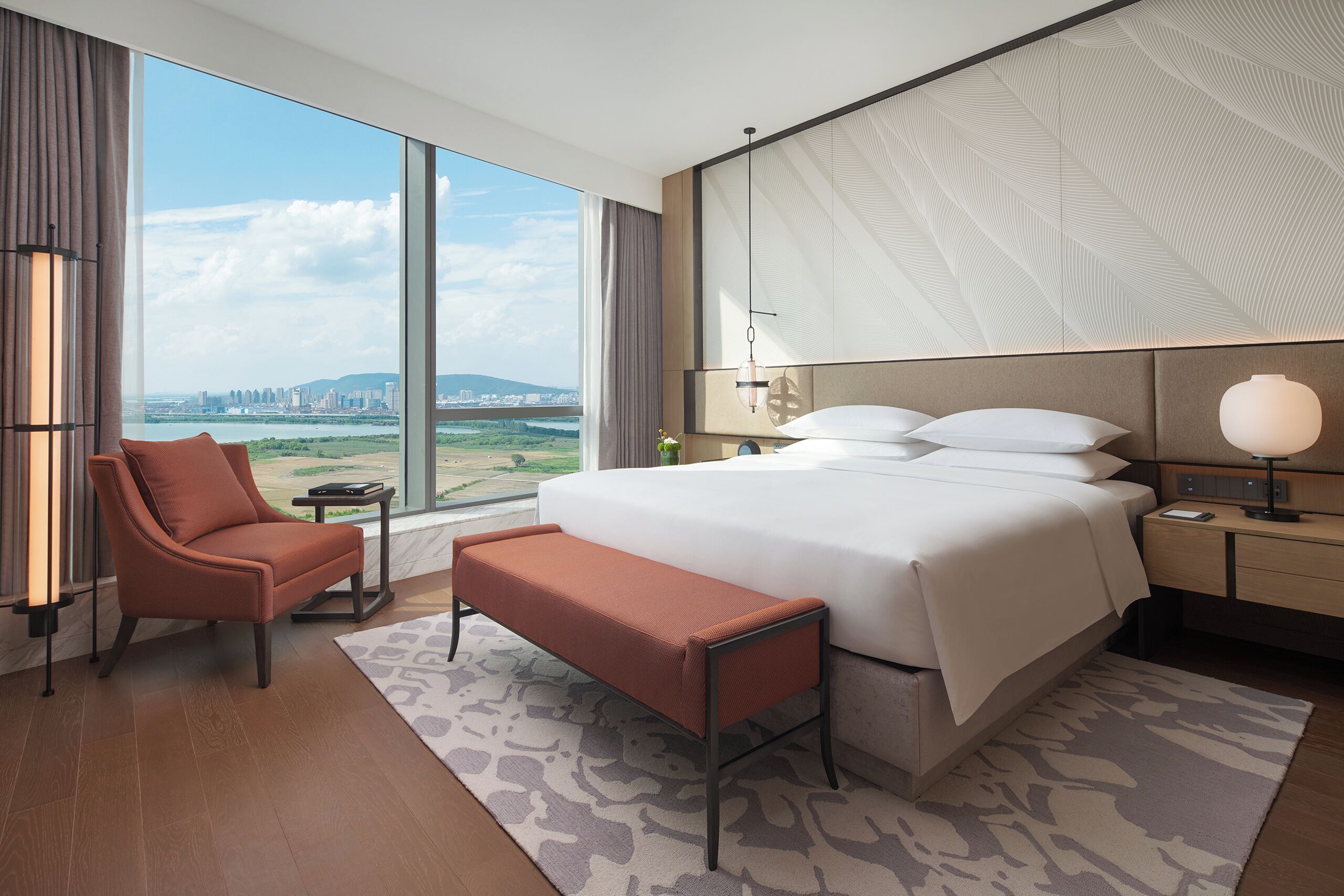 A modern hotel room with a large bed, a seating area, a desk, and a view of a cityscape.