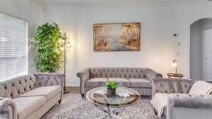 Un salon comprenant un canapé capitonné, une table basse en verre ornée d’une plante et un tableau représentant une scène de forêt.