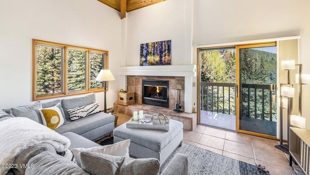 A cozy living room with a fireplace, a sofa, a coffee table, and a large window with a forest view. - Ski In/out with HOT TUB! PENTHOUSE, Walk to Ski School & Restaurants! BC Village (Beaver Creek)