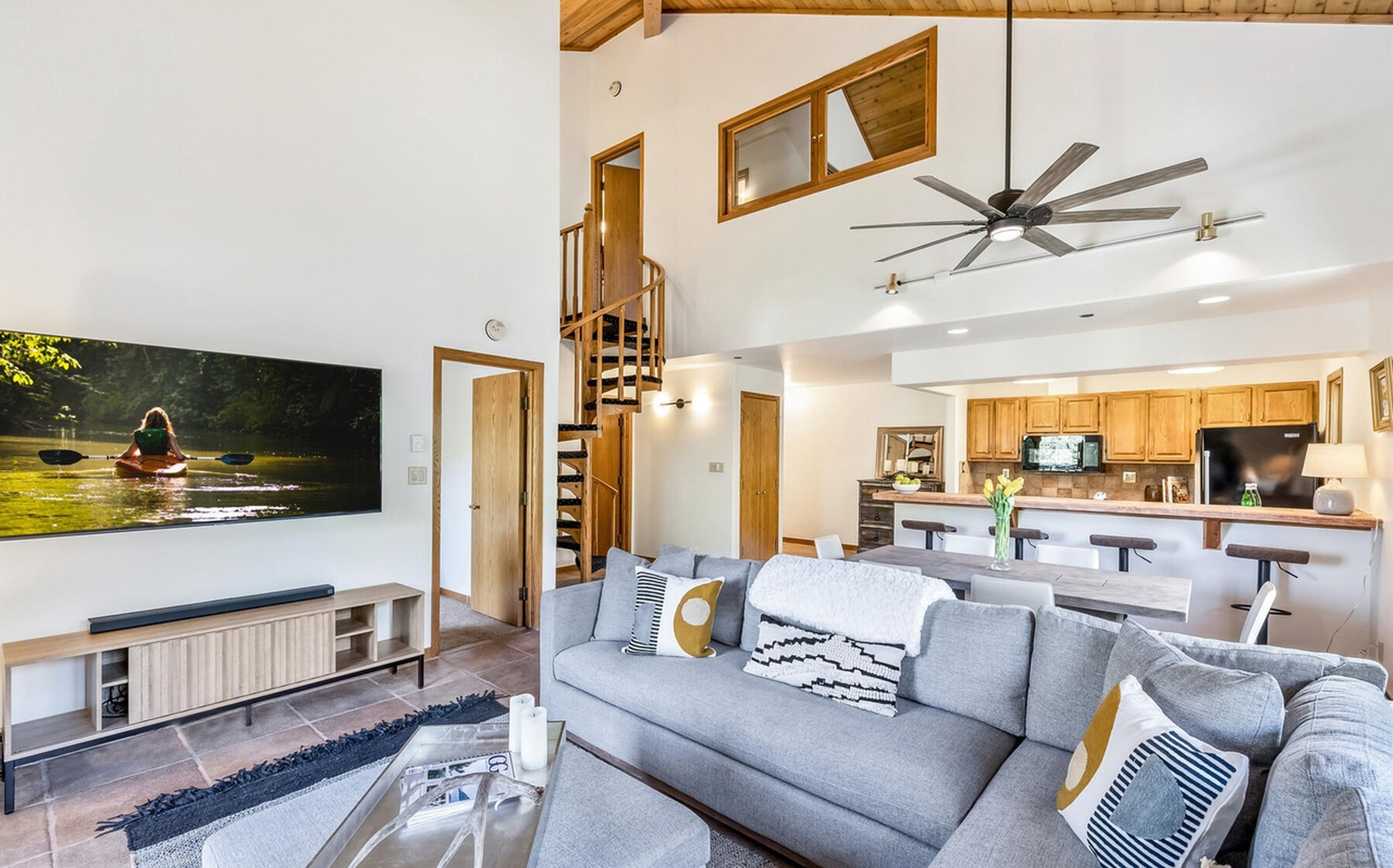 A modern living room with a large flat-screen TV, a grey sofa, and a staircase leading to an upper level.
