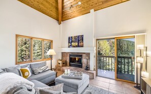 A spacious living room with a fireplace, a sofa set, a dining table, and a balcony with a view of trees.