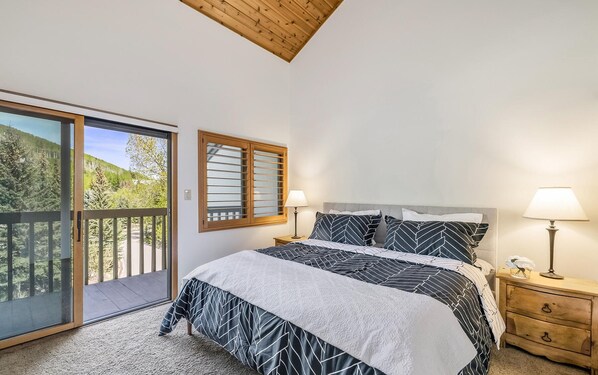 A bedroom with a bed, two bedside lamps, a nightstand, a window with blinds, and a view of a forest.