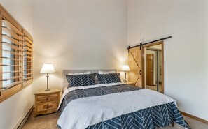 A bedroom with a bed, two bedside tables, a wooden wardrobe, and a window with blinds.