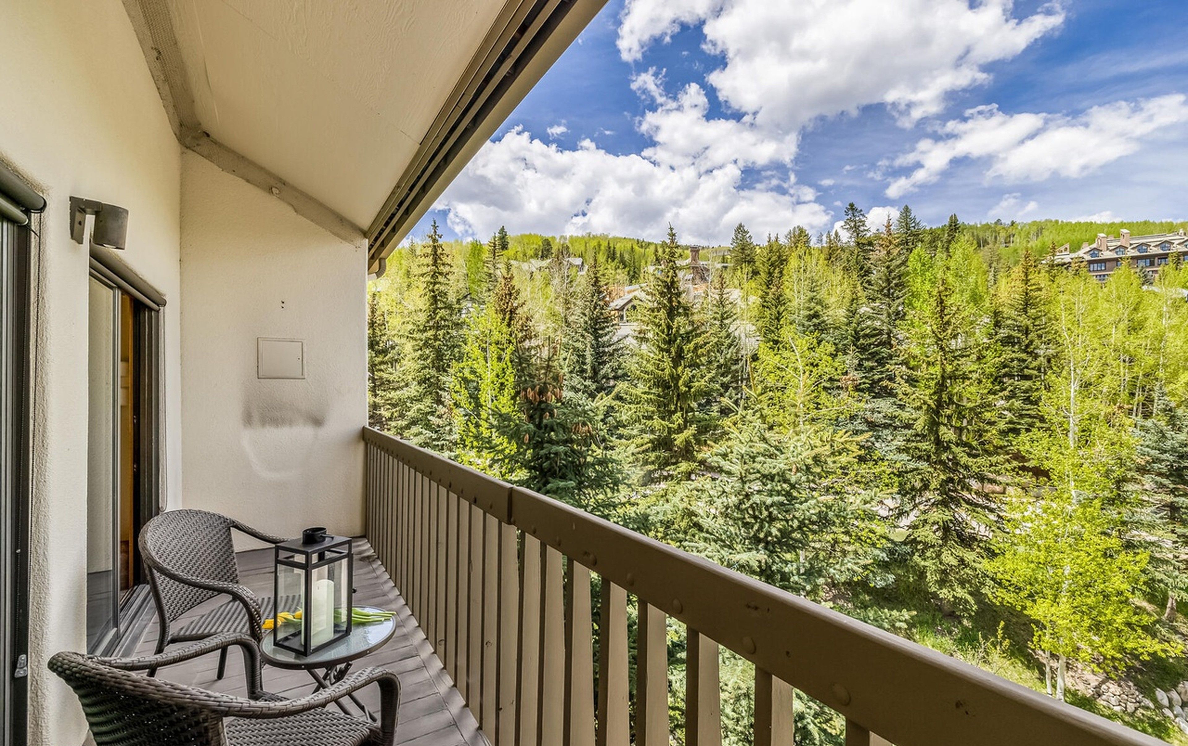 A balcony with wicker chairs and a table, overlooking a forested area with a clear sky.