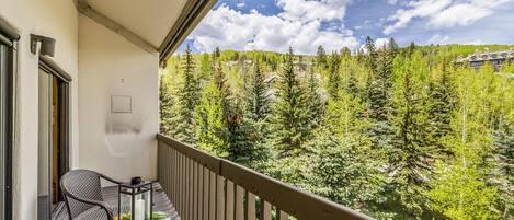A balcony with wicker chairs and a table, overlooking a forested area with a clear sky.