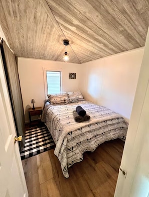 A bedroom with a wooden ceiling, a bed with patterned bedding, a bedside table with a lamp, and a checkered rug.