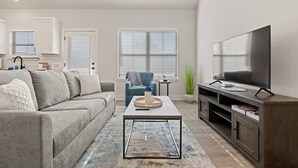 Un salon moderne avec un canapé gris, un fauteuil bleu, une table basse, un téléviseur et, au fond, un coin cuisine.
