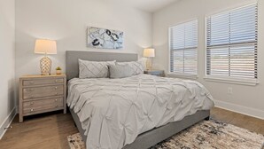 Une chambre à coucher avec un lit, deux tables de chevet, un tableau et deux fenêtres équipées de stores.