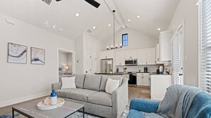 Un vaste espace de vie comprenant un canapé gris, un fauteuil bleu et une table basse. On aperçoit une cuisine à l’arrière-plan, avec des meubles blancs et des appareils électroménagers modernes.