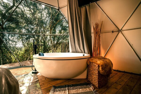A room with a large bathtub, a wooden floor, and a view of trees through a geometric window structure.