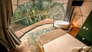 A bedroom with a large bed, a bathtub, and a view of a forest through a geometric window. - Esphera Glamping - Gravataí - RS (Gravatai)