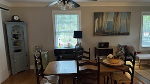 A dining area with a wooden table and chairs, a ceiling fan, a window with blinds, and a clock on a cabinet. - Cozy private efficiency located near downtown Raleigh, PNC Arena, NCSU, RTP, RDU (Raleigh)