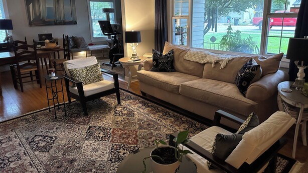A living room with a beige sofa, a patterned rug, a dining area with a table and chairs, and large windows with a view of the outside. - Cozy private efficiency located near downtown Raleigh, PNC Arena, NCSU, RTP, RDU (Raleigh)