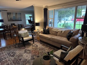 A living room with a beige sofa, a patterned rug, a dining area with a table and chairs, and large windows with a view of the outside.