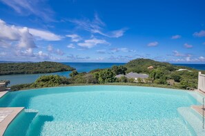 Una piscina d'aigua cristal·lina i de color blau que sembla infinita, amb vista a un paisatge tropical i al mar.