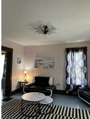 Een moderne woonkamer met een zwart-wit gestreept tapijt, een ronde salontafel, een bank en een stoel. Er hangt een schilderij aan de muur en aan het plafond een kroonluchter.