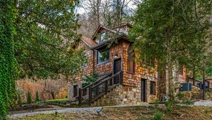 A rustic stone house with a wooden staircase, surrounded by dense trees and a garden. - Stone Cabin Retreat - Dog Friendly - Only 1.5mi to town & access to free trolley (Gatlinburg)