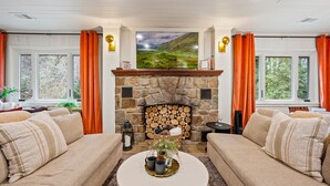 A cozy living room with a stone fireplace, two sofas, a coffee table with decorative items, and large windows with orange curtains. - Stone Cabin Retreat - Dog Friendly - Only 1.5mi to town & access to free trolley (Gatlinburg)