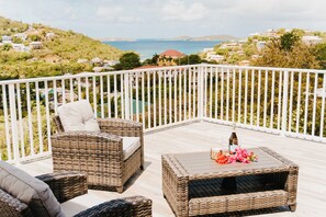A balcony with wicker furniture, a table with flowers and a bottle, and a view of the sea and houses. - Cozy accommodations in a great location. (Cruz Bay)